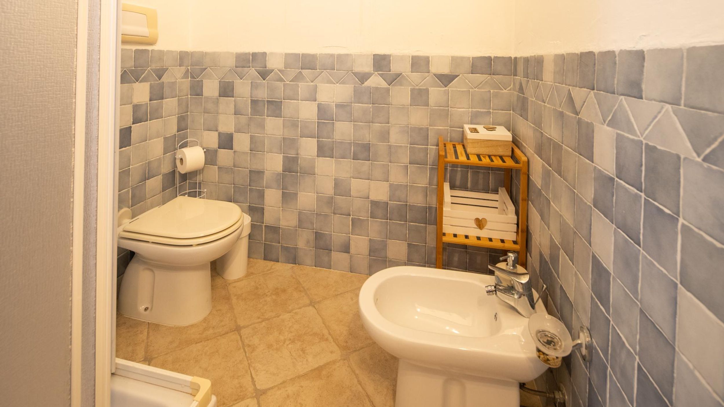 A bathroom with a toilet, bidet, and sink, tiled with a geometric pattern.