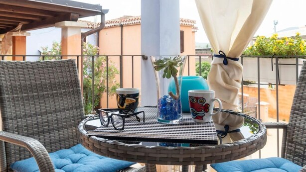 Ein runder Tisch mit einer Glaskaraffe, einer Tasse und zwei Gläsern auf einem Balkon mit Korbstühlen und Blick auf Gebäude.
