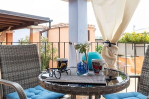 A round table with a glass vase, a mug, and a pair of glasses on a balcony with wicker chairs and a view of buildings. - Welcomely - Il Nido Blu (Tortoli)