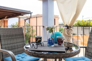 Una mesa redonda con un jarrón de vidrio, una taza y un par de gafas en un balcón con sillas de mimbre y vistas a edificios.