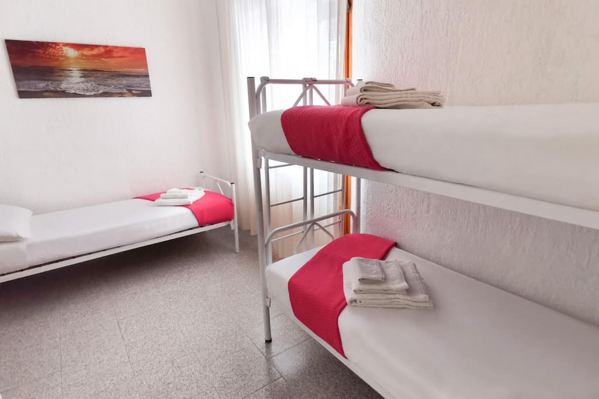 A dormitory room with bunk beds, a painting of a sunset, and a window with curtains.