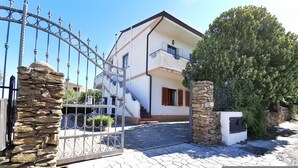A two-story house with a stone gate, a paved driveway, and a large tree in front. - Welcomely - La Casa Di Rita - Salamaghe (Budoni)