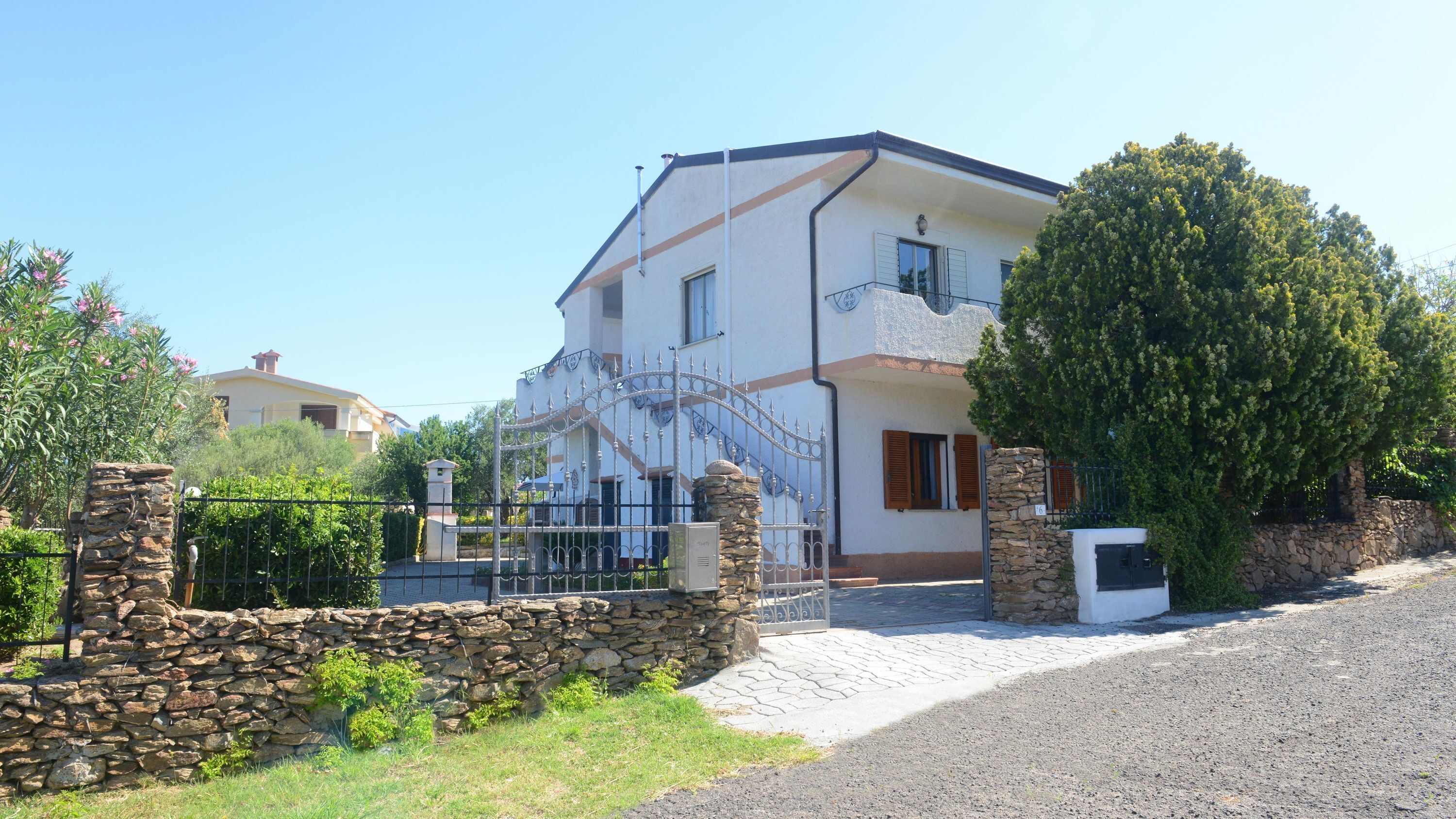A two-story house with a stone wall entrance, a metal gate, and a large tree in front.