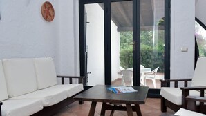 Uma área de pátio com um sofá branco, uma mesa de madeira e duas cadeiras. Uma porta de vidro dá para o exterior.