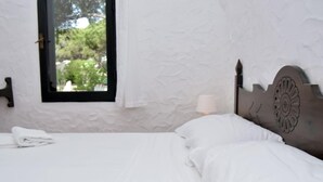 Cama bem arrumada com lençóis brancos num quarto com parede branca texturizada e uma janela.