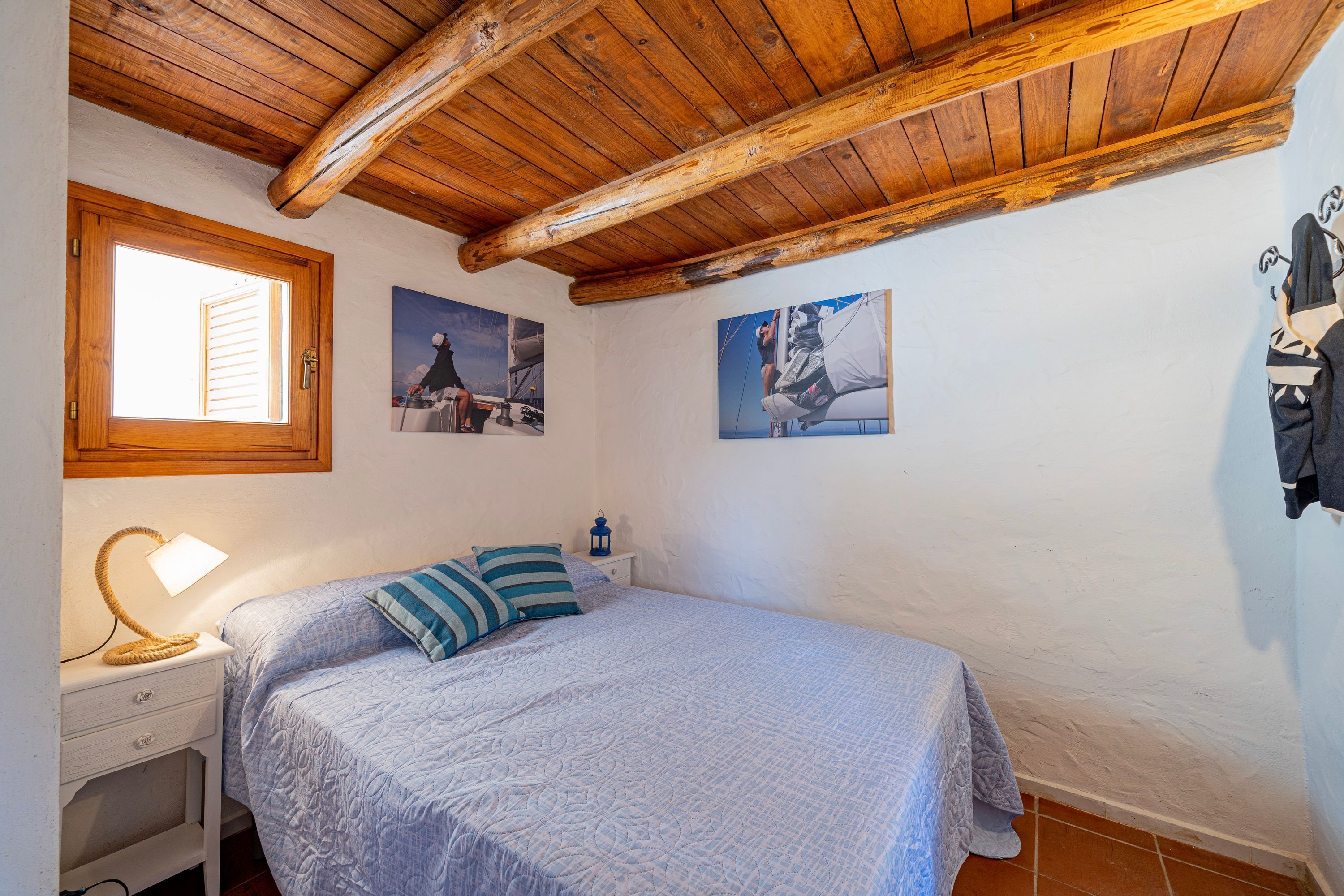 Ein Schlafzimmer mit Holzboden, einem Bett mit blau-weißer Bettdecke, einem Nachttisch mit Lampe und einem Fenster mit Jalousien.