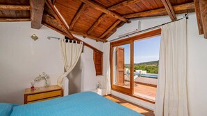 A bedroom with a wooden ceiling, a bed with a blue bedspread, a nightstand, a window with white curtains, and a view of a pool and greenery outside. - Welcomely - Le Vele in Olbia (Olbia)