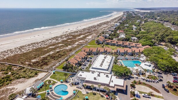 An aerial view of a coastal resort with a beach, swimming pool, and multiple buildings.