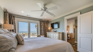 A spacious bedroom with a large bed, a ceiling fan, a TV, and a view of the beach.