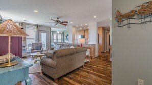 A spacious living area with a sofa, a coffee table, and a lamp. There is a kitchen in the background with wooden cabinets and a dining area.