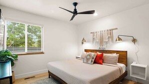Een slaapkamer met een bed, een plafondventilator, een nachtkastje met lamp, een raam met jaloezieën en een plant.