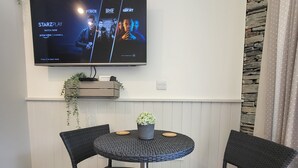 Moderni olohuone, jonka seinälle on asennettu litteänäyttöinen televisio, ja jonka ruudulla näkyy elokuvan juliste. Huoneessa on pieni pöytä, jonka ympärillä on kaksi tuolia, ja pöydällä on ruukkukasvi.