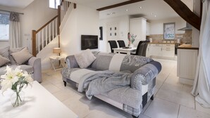 En moderne stue med en grå sofa, et hvidt sofabord og en vase med hvide blomster. Der er en trappe til et andet niveau, et spiseområde med stole, og et køkken med hvide skabe.