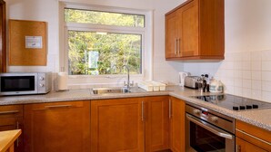 Cocina con gabinetes de madera, microondas, lavamanos, estufa y ventana con vistas al verdor.