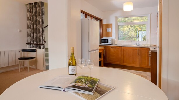 Una cocina moderna con un refrigerador blanco, microondas y armarios de madera. Se ve una mesa de comedor con una botella de vino y copas.