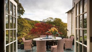 Eine Terrasse mit rundem Tisch und Korbstühlen, umgeben von Topfpflanzen und Blick auf Herbstlaub.