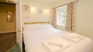 A neatly made bed with white linens and towels, a wooden headboard, a small shelf with decorative items, and a window with floral curtains. - 2 Bridge Syke, Elterwater (Ambleside)