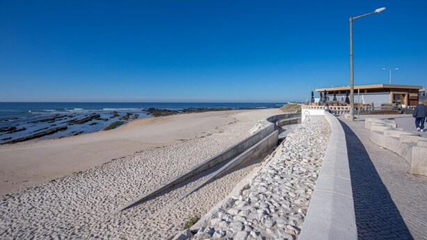 A beach with a stone walkway, a building with a flat roof, and a clear blue sky.