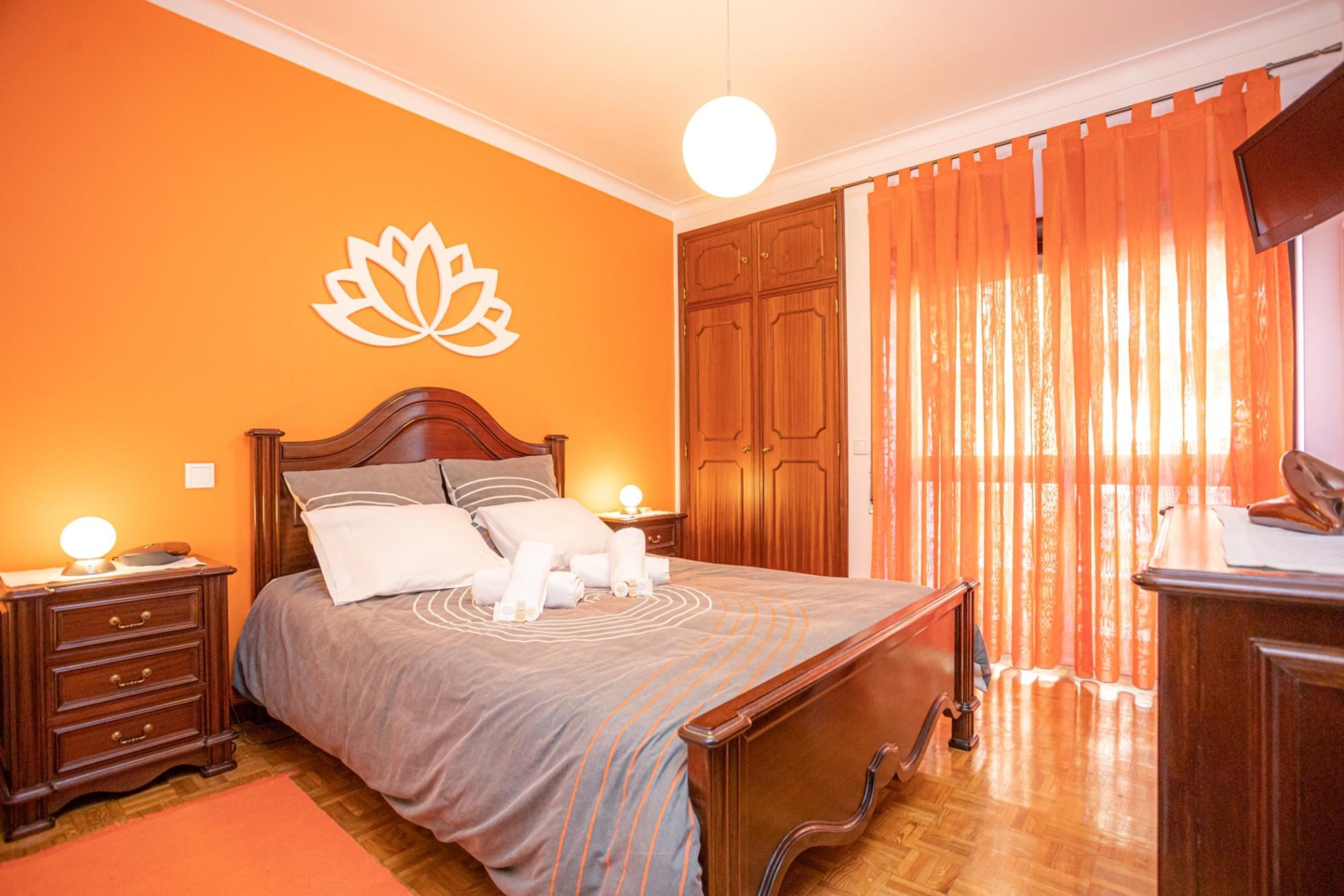 A bedroom with a wooden bed, a nightstand with a lamp, a wall-mounted TV, and a large window with orange curtains.