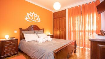 A bedroom with a wooden bed, a nightstand with a lamp, a wall-mounted TV, and a large window with orange curtains.