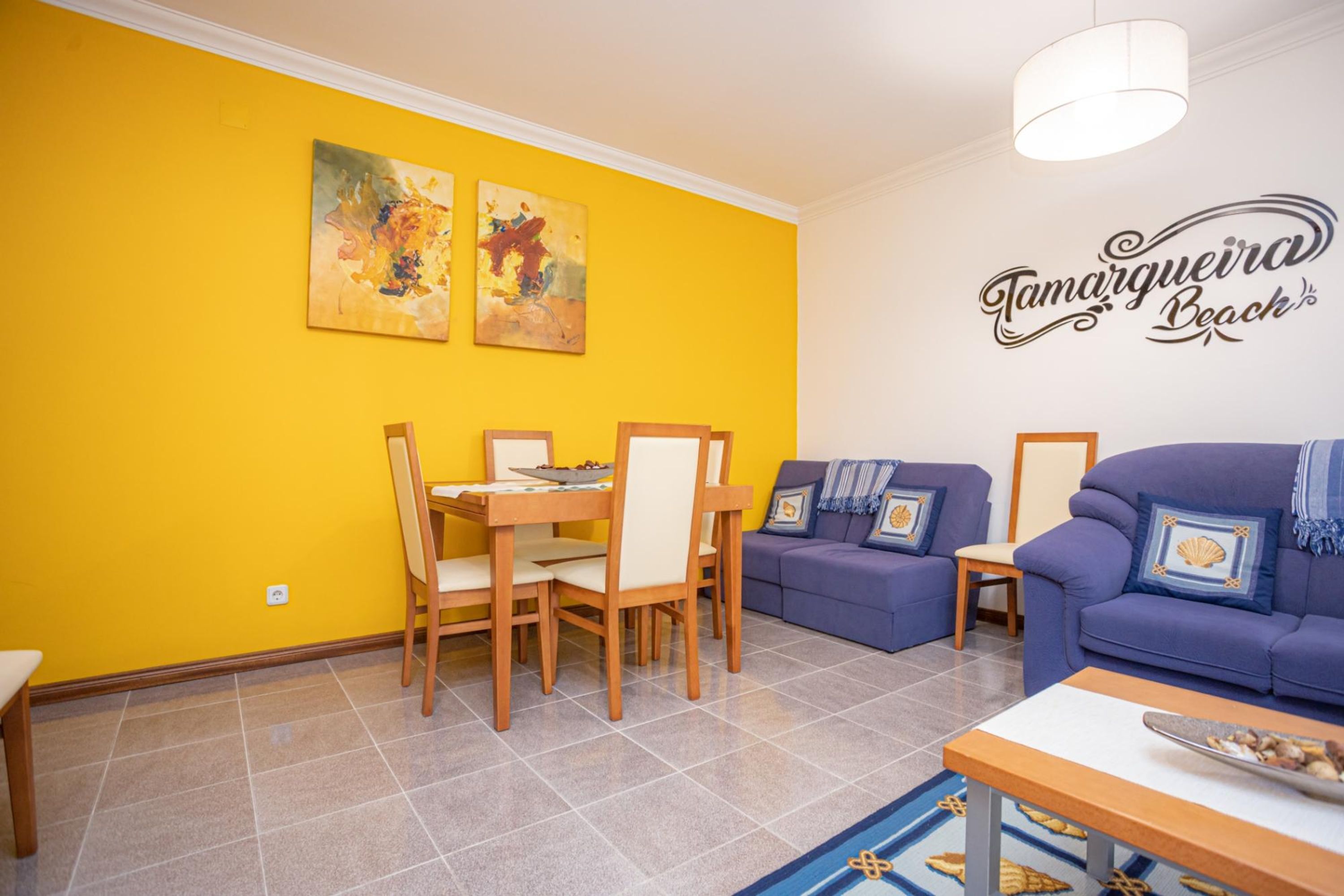 A bright yellow room with a dining table, two sofas, and a wall sign reading "Tamarqueira Beach".