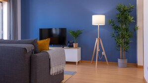 Een moderne woonkamer met een blauwe accentmuur, een flatscreen televisie, een staande lamp, een potplant en een grijze bank.