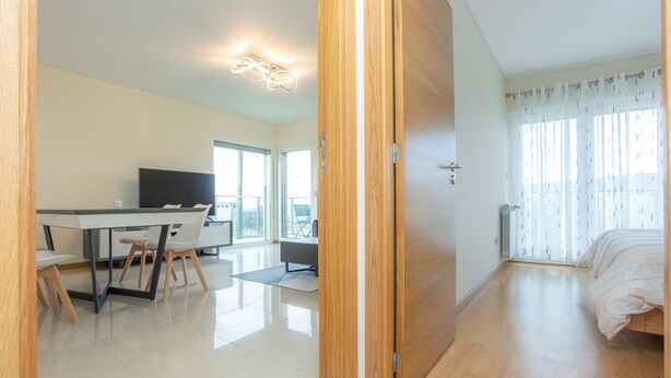 Una habitación de hotel moderna con puerta de madera, cama, escritorio con sillas, televisor y ventanas grandes.