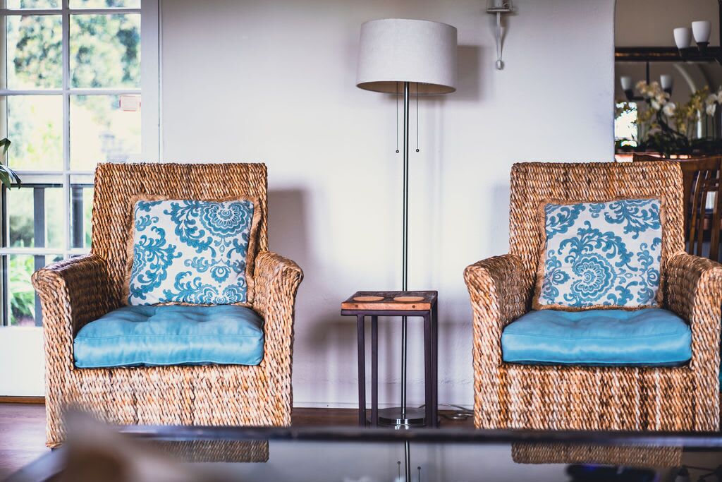 Two wicker armchairs with blue cushions and a small side table, a floor lamp, and a view of greenery outside.