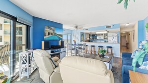 A modern living room with a sofa, a dining table, and a kitchen area with a bar. - Crescent Towers II 608 - Oceanfront 2 BR Condo (North Myrtle Beach)