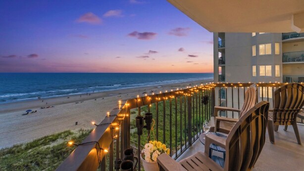 A balcony with a view of the beach, outdoor seating, and string lights. - Crescent Towers II 608 - Oceanfront 2 BR Condo (North Myrtle Beach)