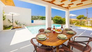Un espacio al aire libre cubierto con área de comedor, piscina, mesa de madera, sillas de mimbre y sombrillas.