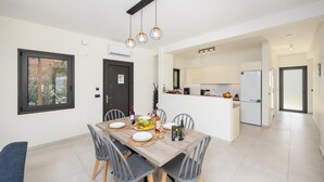 A modern kitchen with white cabinets, a built-in oven, and a dining area with a wooden table and chairs. - Villa Petra - Two Bedroom Villa, Sleeps 4 (Afrato)