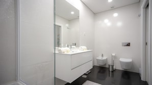 Un baño moderno con cabina de ducha de vidrio, tocador blanco con dos lavamanos, inodoro y bidé.