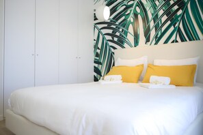 Un dormitorio con cama, paredes blancas, un mural de hojas tropicales y un armario blanco.