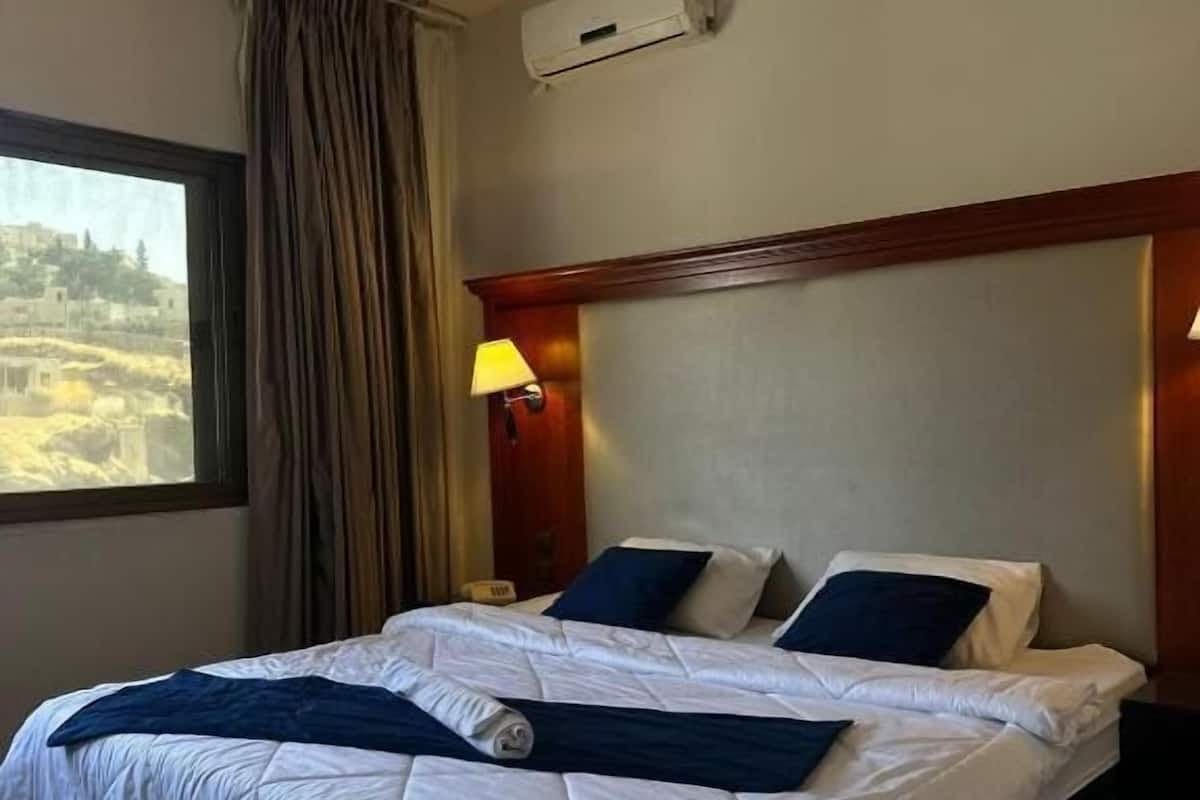 A hotel room with a large bed, a window with curtains, an air conditioning unit, and a nightstand with a lamp.