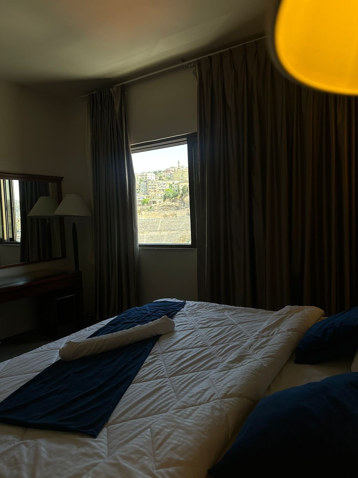 Een hotelkamer met een bed, een raam met gordijnen, een bureau met een lamp en uitzicht op een stadsbeeld.