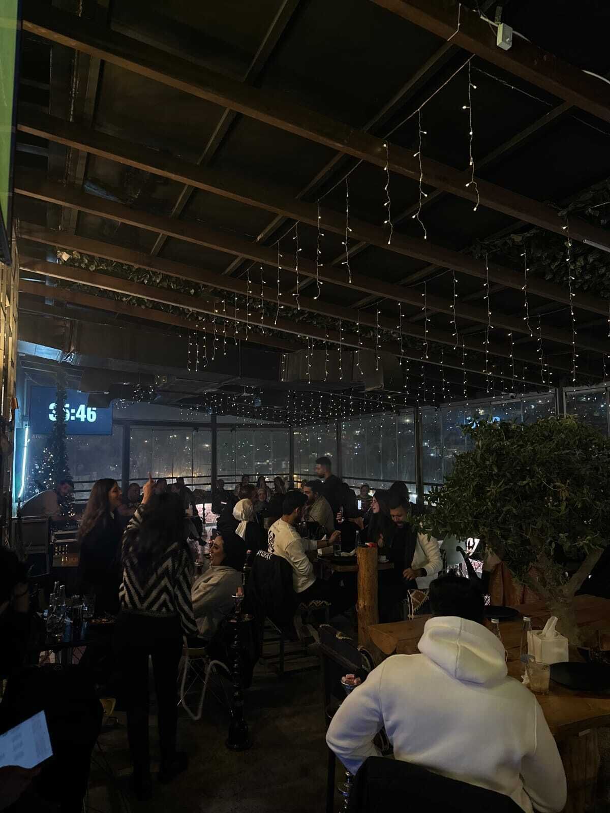 Un bar situé sur un toit, décoré de guirlandes lumineuses, avec une horloge indiquant 3h46, et offrant une vue sur le paysage urbain.