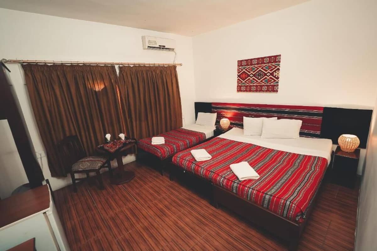 A hotel room with two beds, a small table, a chair, a lamp, and a decorative wall hanging.
