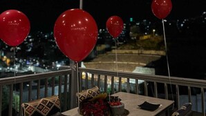 Un balcon avec une table dressée pour deux personnes, des ballons rouges et une vue sur la ville illuminée la nuit.