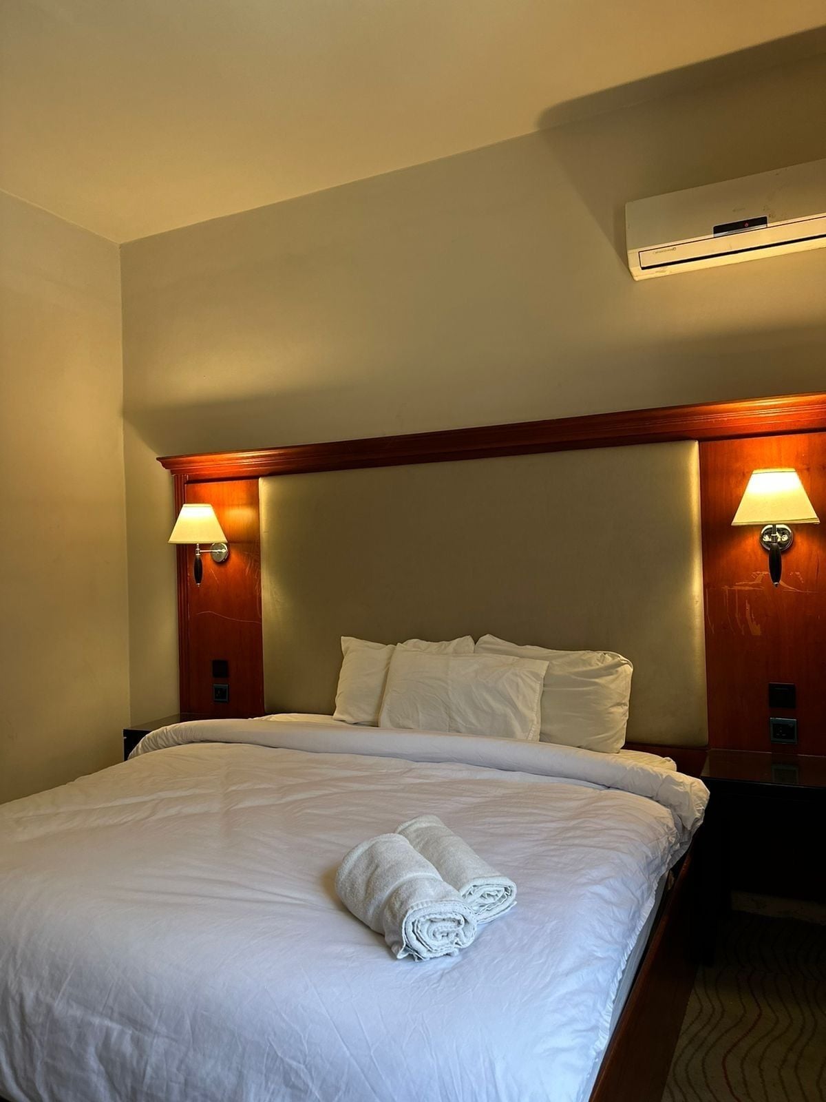 A hotel room with a large bed, two bedside lamps, an air conditioning unit, and a wooden headboard.