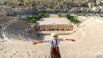 An ancient amphitheater with tiered seating and a central stage, surrounded by a hilly urban area with buildings.