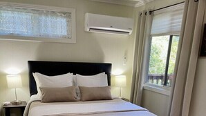 A neatly arranged bedroom with a bed, bedside lamps, a window with curtains, an air conditioning unit, and a decorative ottoman.