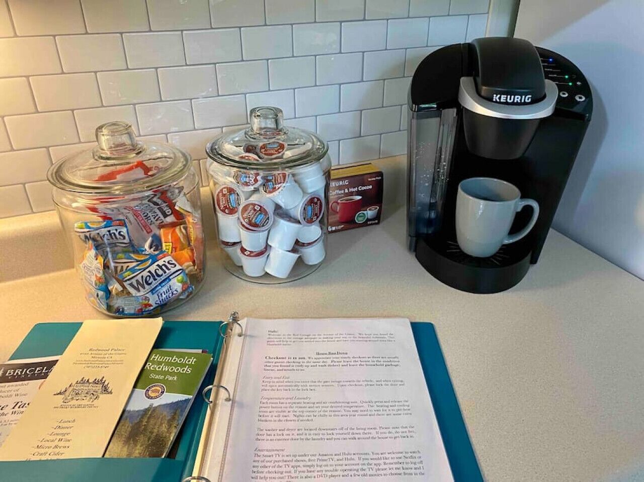 A kitchen counter with a Keurig coffee maker, two containers of coffee pods, a mug, and a travel guide.