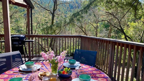 A round table set for a meal on a wooden deck with a view of trees and mountains.