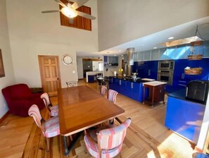 Un vaste espace de salle à manger avec une table en bois et des chaises à rayures, un fauteuil rouge et une cuisine dotée de meubles bleus et d’appareils électroménagers modernes.