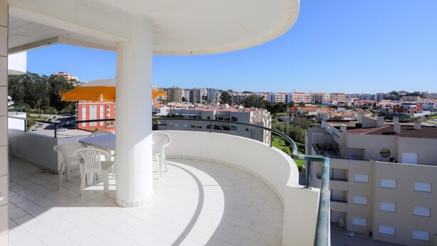 Un balcón con sillas y una mesa blancos, con vista a un paisaje urbano con edificios y vegetación.