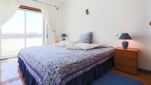 Un dormitorio con una cama grande, dos mesitas de noche con lámparas, un aparador de madera y vistas a través de un ventanal.