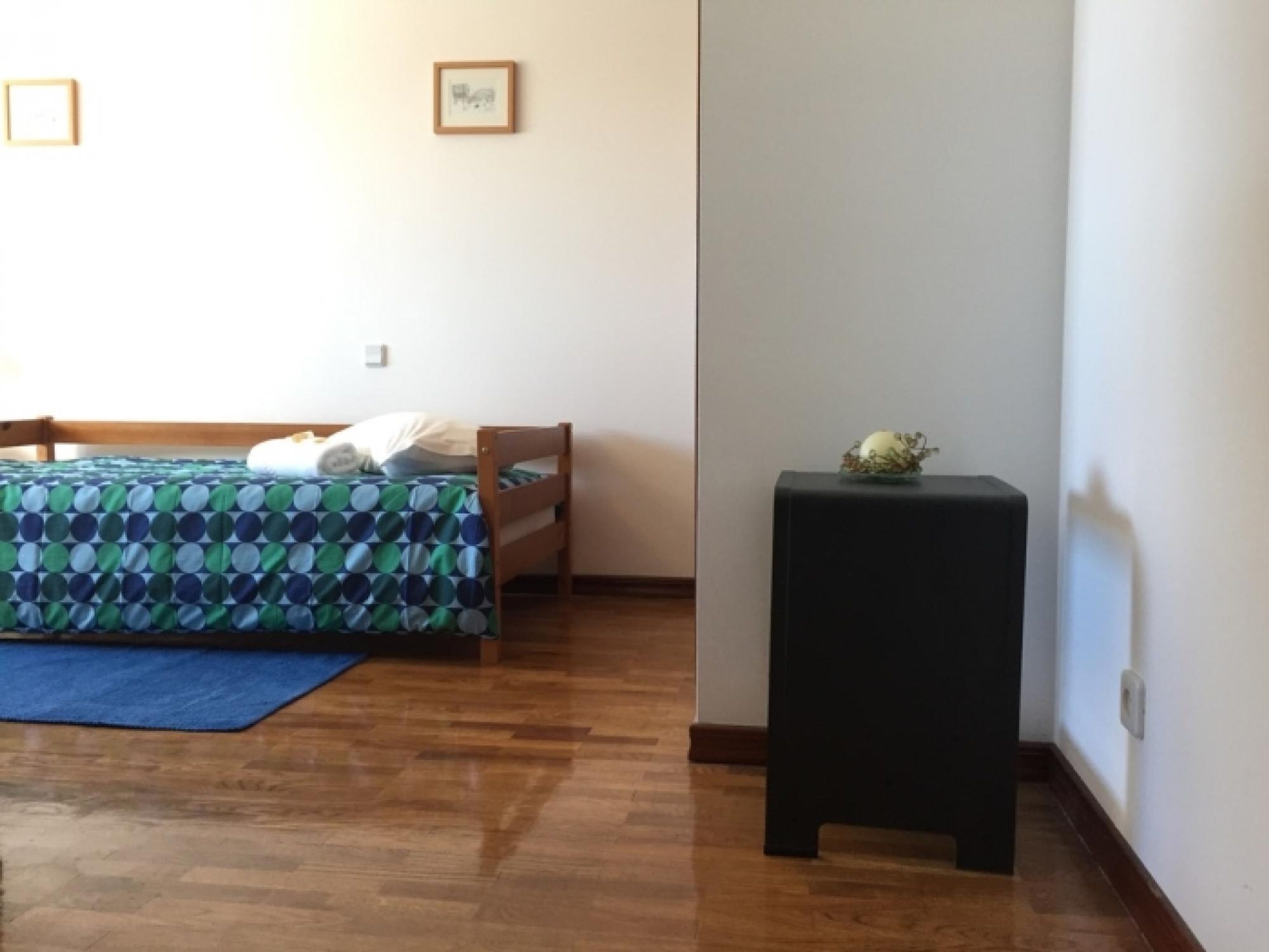 A bedroom with a bed, a blue rug, a small table with a plant, and a framed picture on the wall.