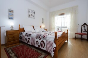 Ein Schlafzimmer mit einem hölzernen Bett, einem roten Teppich mit Muster, einem Holzstuhl und einem Fenster mit durchsichtigen Vorhängen.