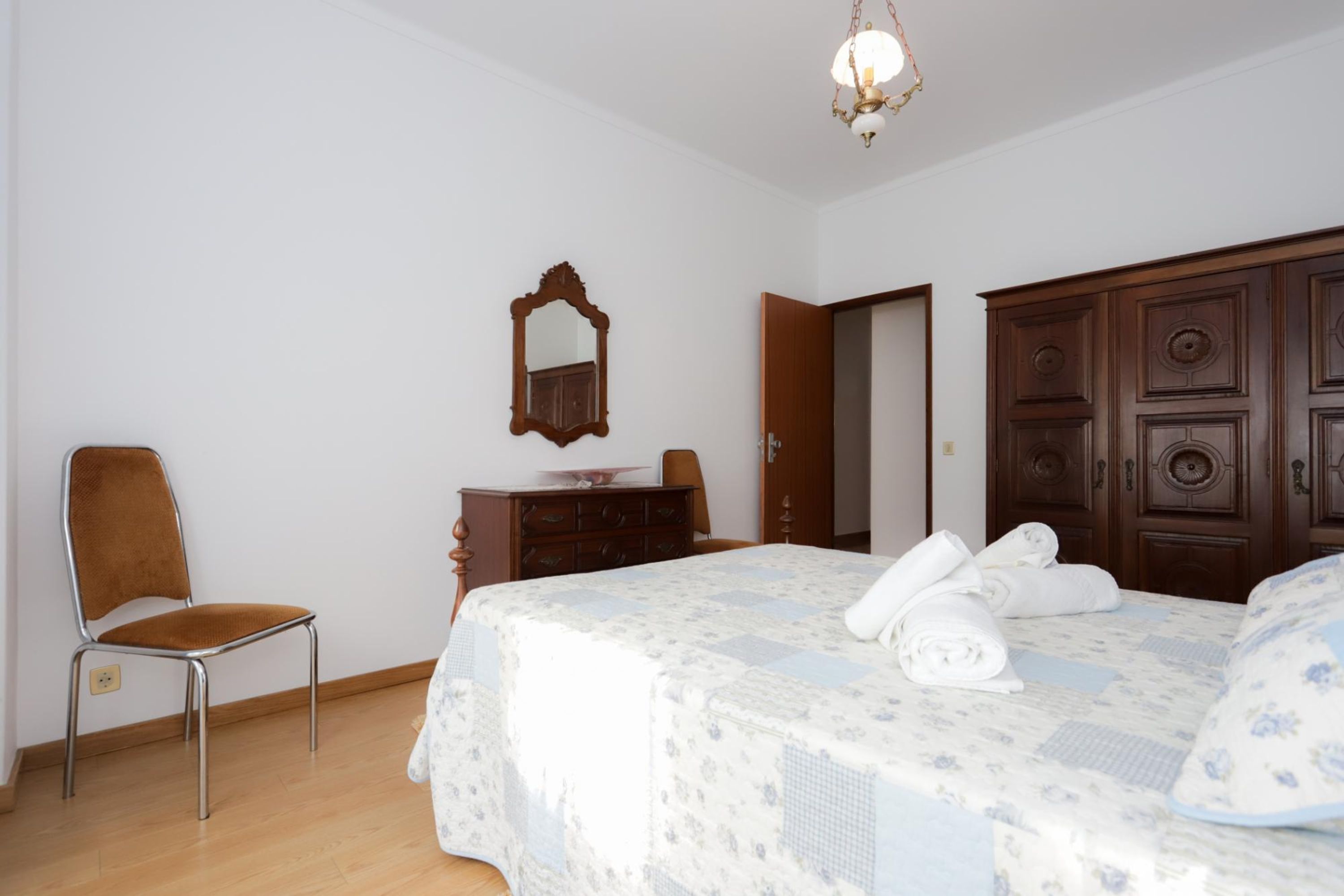 A bedroom with a bed, a wooden wardrobe, a chair, a dresser, and a mirror.